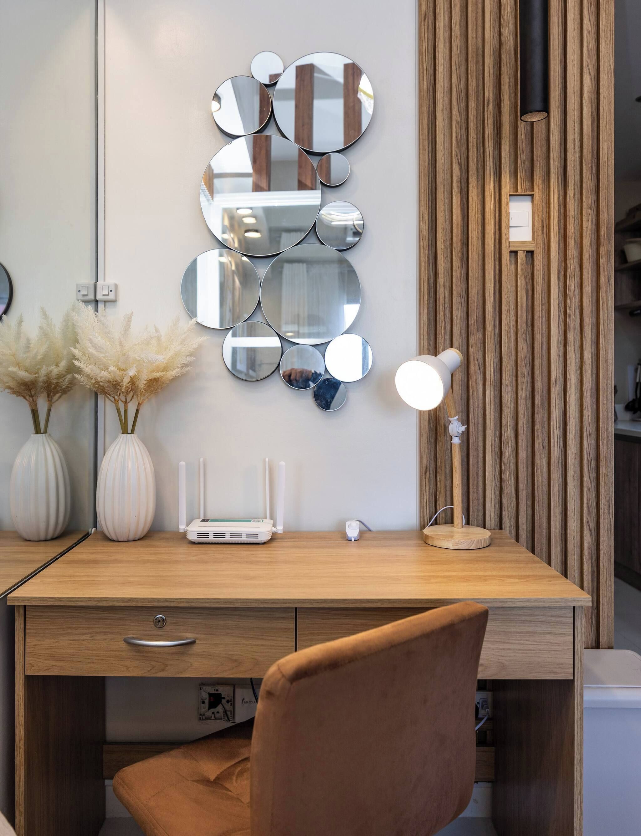 Ein moderner Schreibtisch mit Holzoptik, eine weiße Lampe und eine Wandanordnung von Spiegeln.