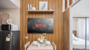 A modern room with a wooden wall panel, a flat-screen TV mounted above a dining table with white plates and cups, a microwave, and a refrigerator. - Elegant Wood-accent Studio With Balcony (Lapu-Lapu)