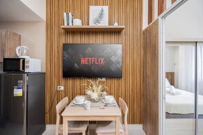 A modern room with a wooden wall panel, a flat-screen TV mounted above a dining table with white plates and cups, a microwave, and a refrigerator. - Elegant Wood-accent Studio With Balcony (Lapu-Lapu)