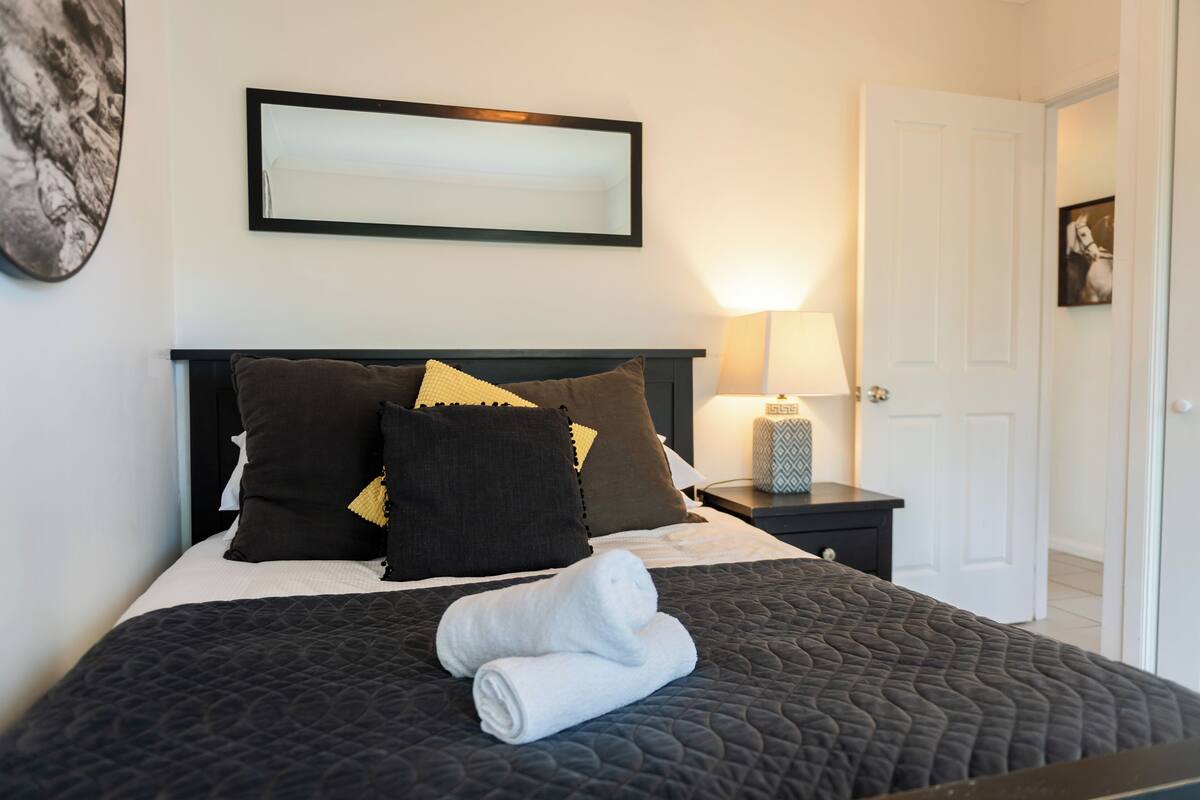 A neatly made bed with a dark quilt, multiple pillows, and a rolled white towel.