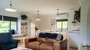 A modern living room with a brown leather sofa, a white coffee table, and a blue armchair. A kitchen with white cabinets and a dining area with white chairs is visible in the background. - The Woods Farm (Tomerong)