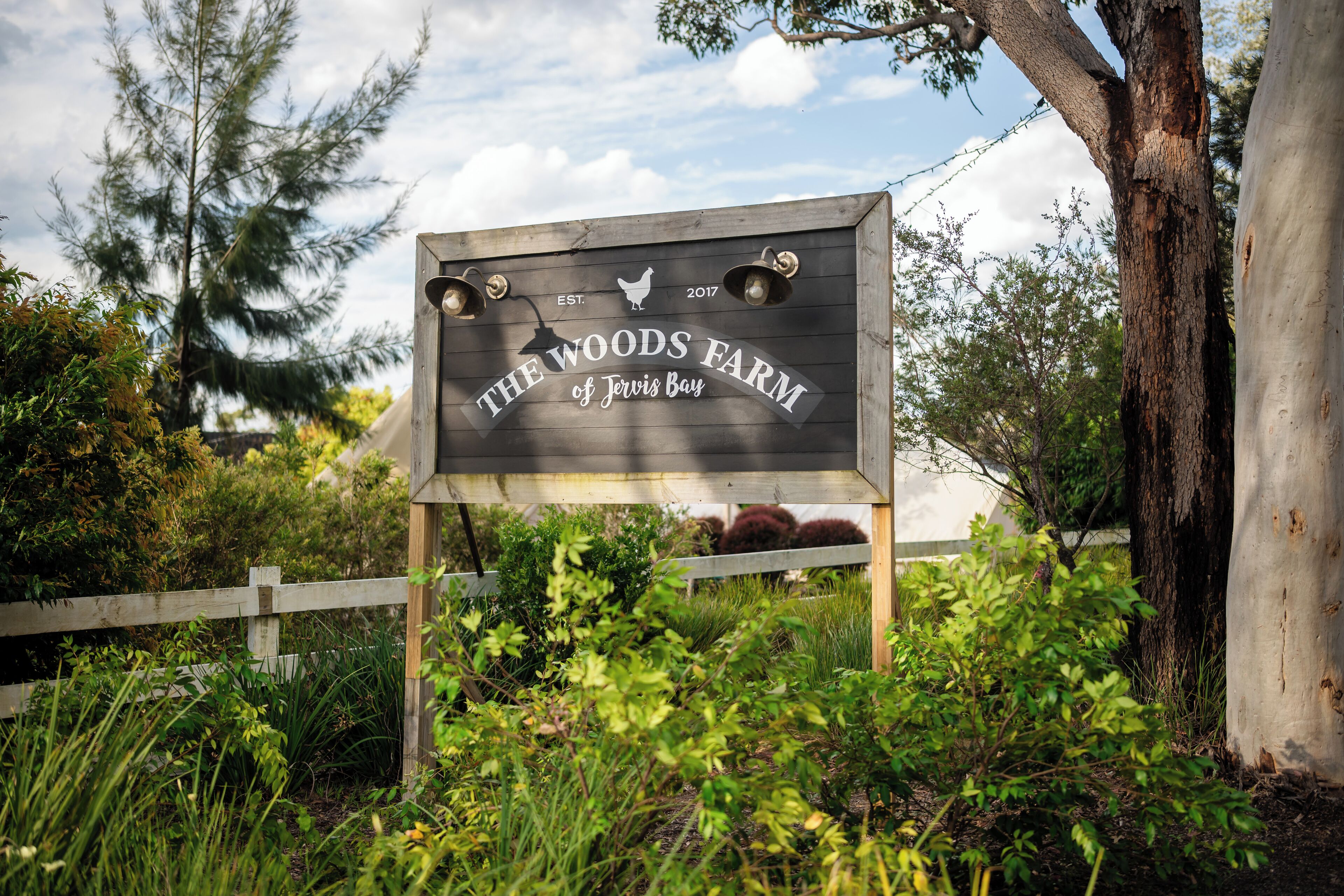 Puinen kyltti, jossa lukee ”The Woods Farm of Jervis Bay” ja on kuvattuna kanan logo. Kylttiä ympäröi vehreää kasvillisuutta ja puita.