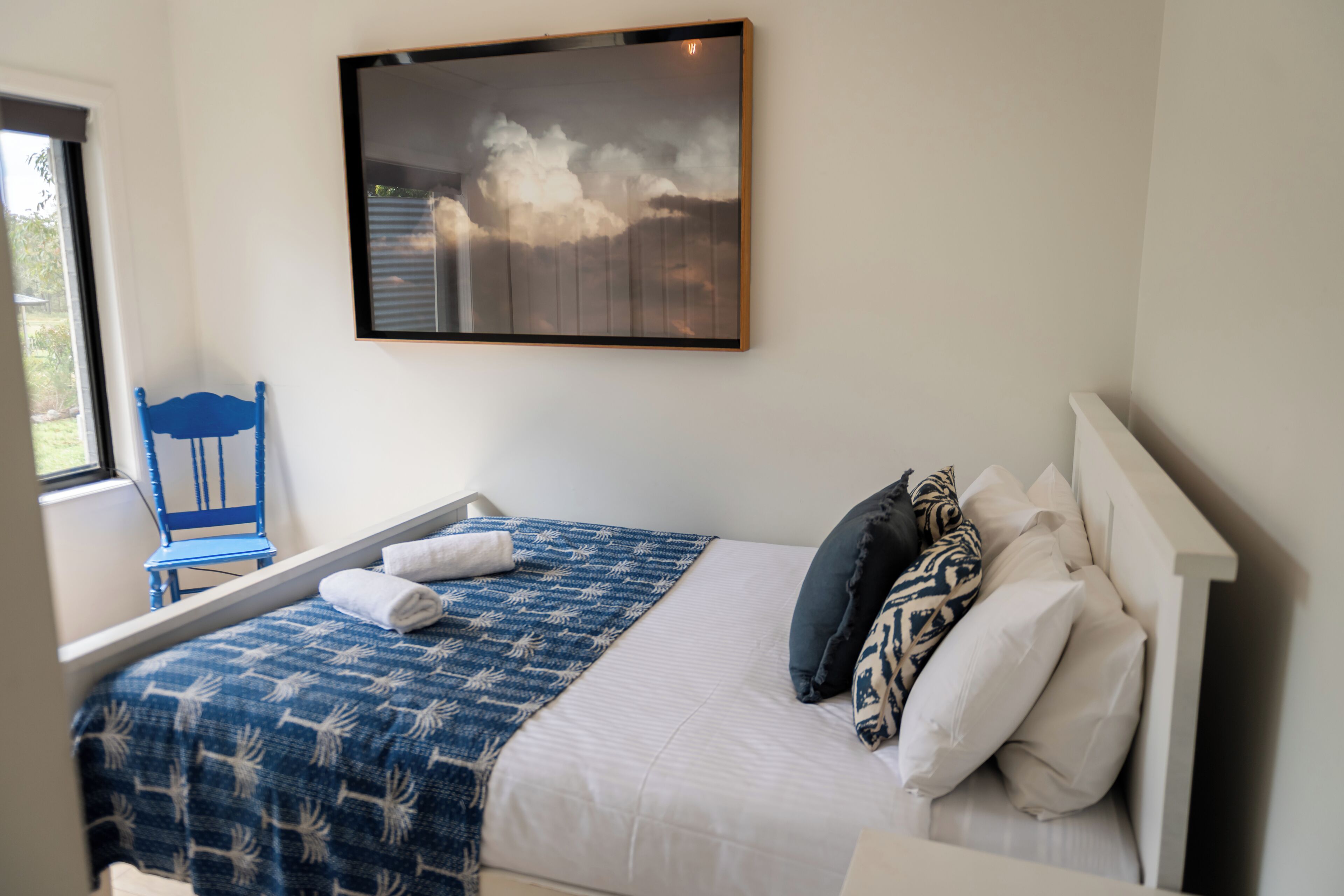 A neatly made bed with a blue patterned bedspread, a white pillow, and a blue chair by the window.