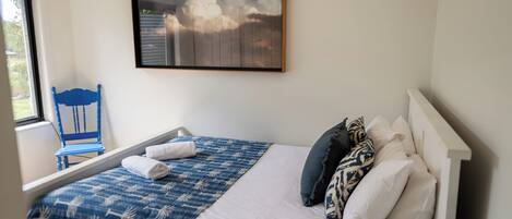 A neatly made bed with a blue patterned bedspread, a white pillow, and a blue chair by the window.