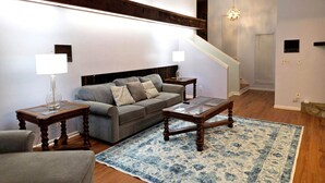 Ein Wohnzimmer mit einer Couch, zwei Beistelltischen, einem Couchtisch, einem blauen Teppich mit Muster und einer Treppe.