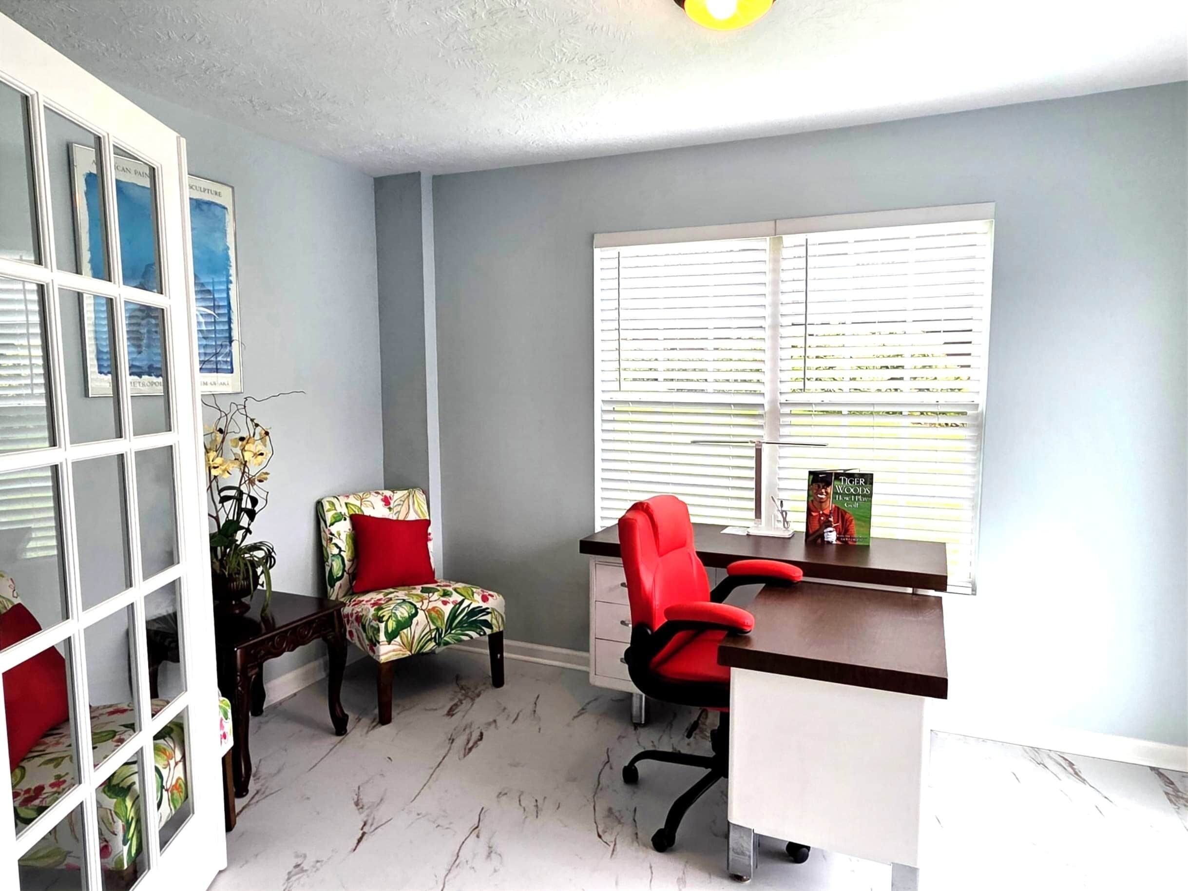 A room with a desk, two chairs, a window with blinds, a plant, and framed artwork on the wall.
