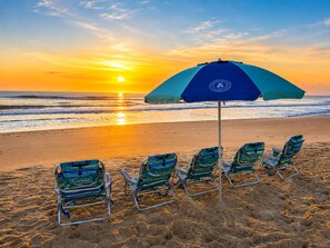 Escena de playa con un atardecer, una sombrilla azul y cuatro sillas de playa a rayas.