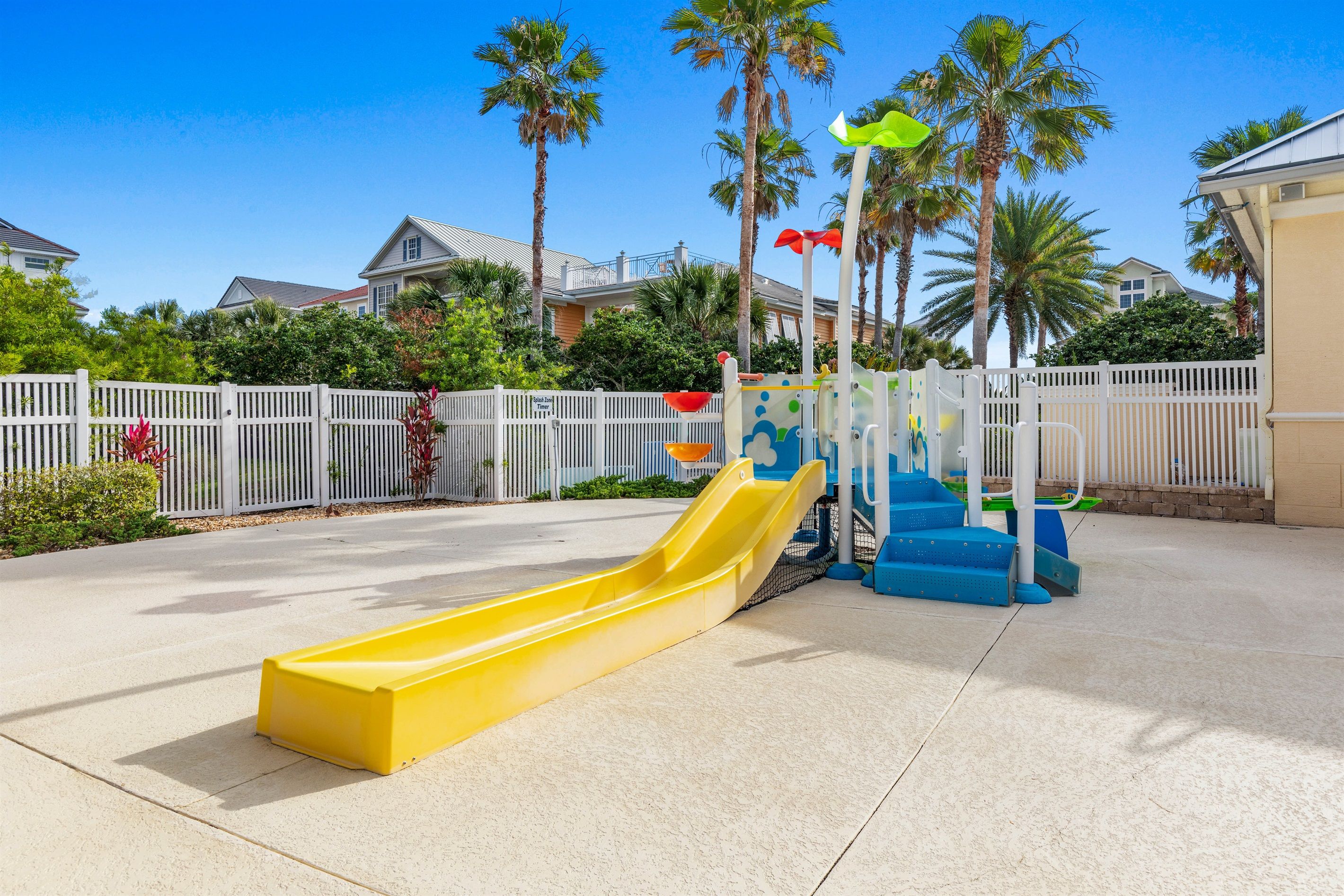 Un parque infantil con un tobogán amarillo y escalones azules, rodeado por una valla blanca y palmeras.