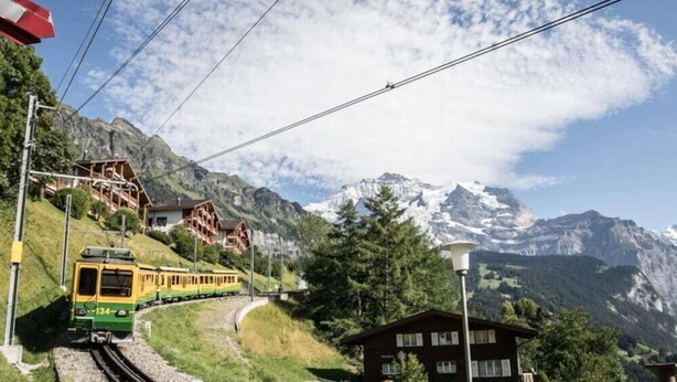 Un tren amarillo sobre rieles, con montañas al fondo y una casa en primer plano.
