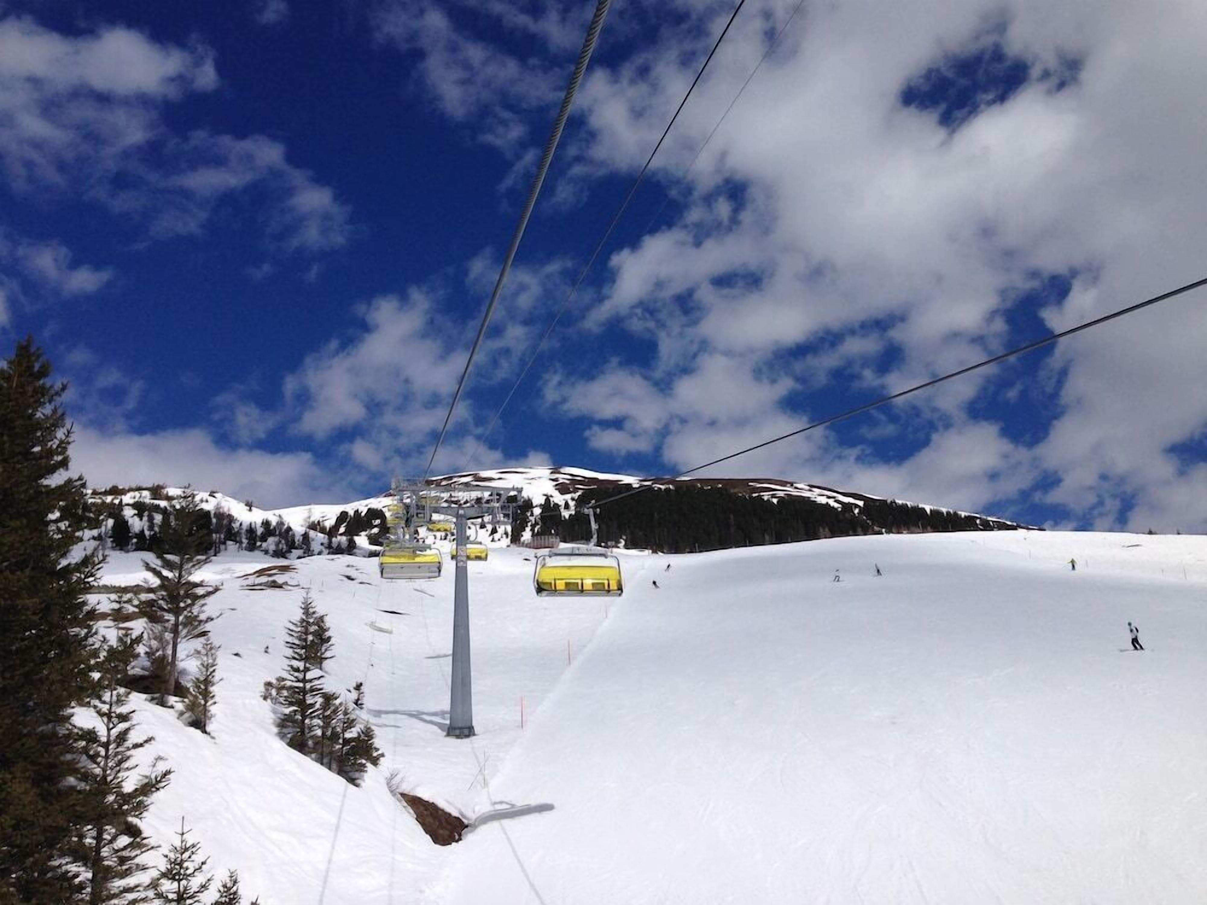 A ski lift with yellow chairs against a snowy mountain backdrop under a partly cloudy sky.