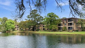 Una vista tranquila de un lago con un edificio de varios pisos, balcones y un césped.