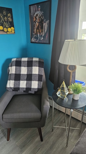 Ein Zimmer mit blauer Wand, einer grau gestreiften Armlehnenschaukel und einer Lampe mit goldfarbener Basis, sowie einem gerahmten Gemälde.
