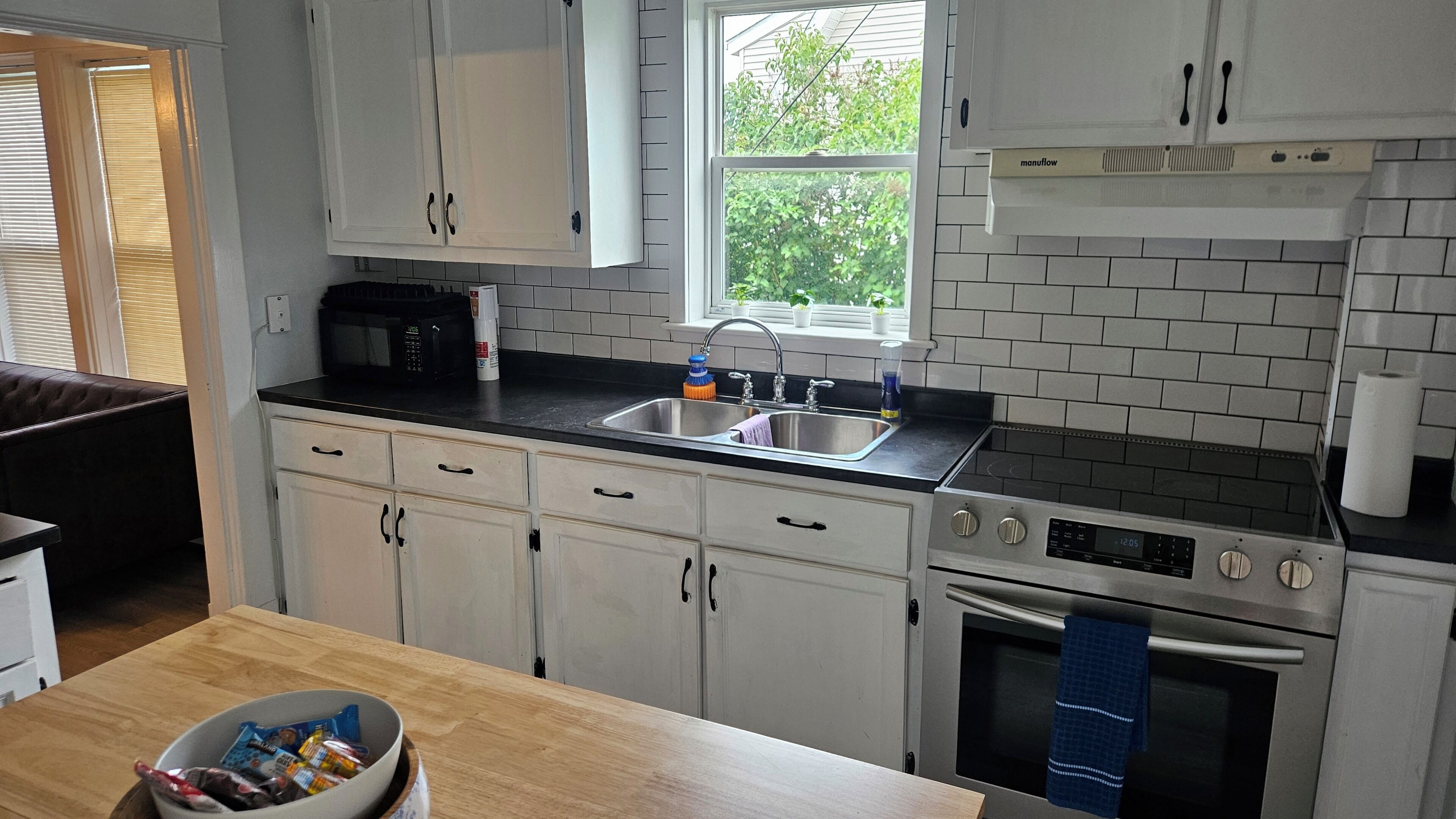 A kitchen with white cabinets, a black countertop, a stainless steel sink, a microwave, a stove, and a window with a view of greenery outside.