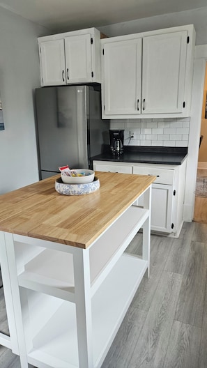 Una cocina con armarios blancos, un refrigerador de acero inoxidable, una cafetera y una mesa de madera con un tazón.