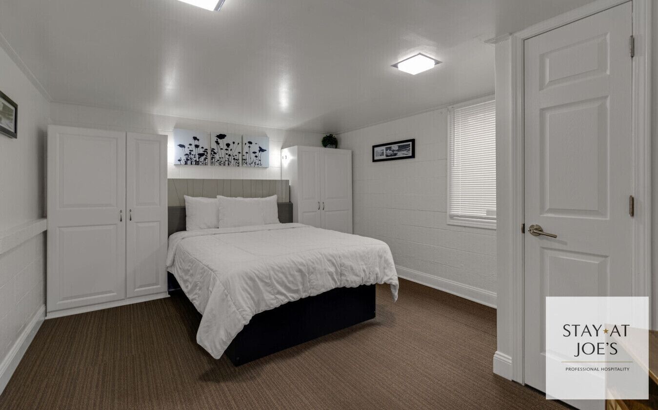 Une chambre à coucher avec un lit, des murs blancs et une porte. On y trouve des armoires et un luminaire au plafond.