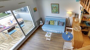 Ein kompakter Wohnbereich mit einem blauen Sofa, einem kleinen Tisch und Blick durch eine Glastür auf einen Außenbereich mit Holzmöbeln.