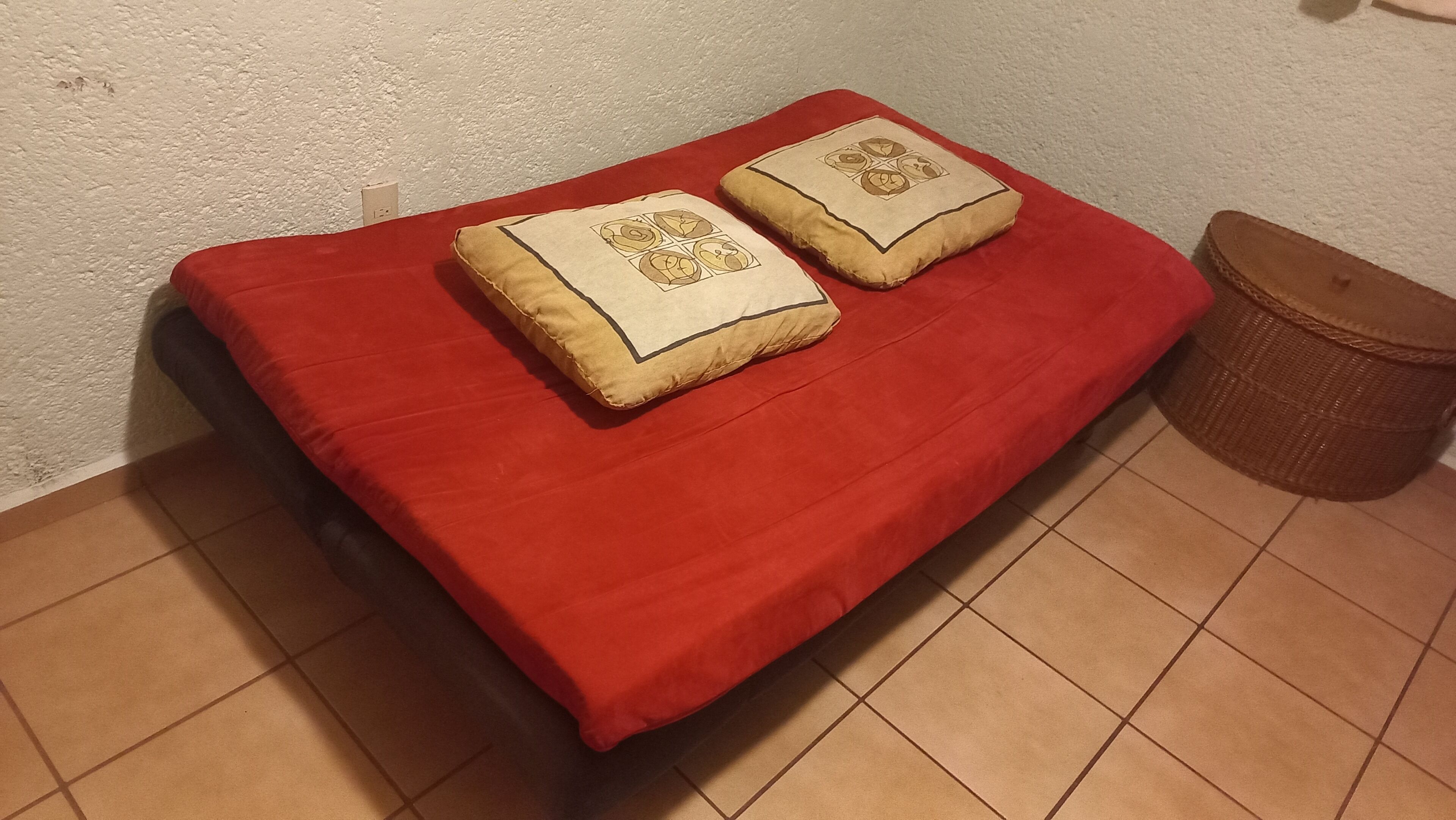 Een rode bank met twee decoratieve kussens tegen een structuurwand, een rieten mand en een betegelde vloer.