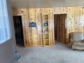 Una habitación con paneles de madera, una estantería, una silla y una puerta que da a otra habitación.