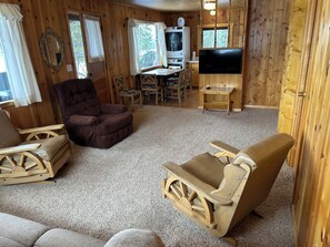 A wooden cabin interior with a sofa, armchair, TV, and a dining area.