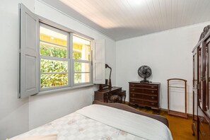 Une chambre à coucher avec un lit, une commode, un bureau, un ventilateur et une fenêtre dont les volets sont ouverts.
