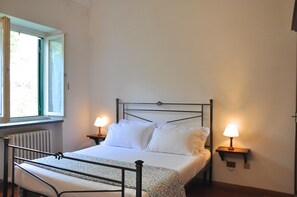 Ein ordentlich bezogenes Bett mit weißen Leinen, zwei Nachttische mit Lampen, ein Fenster mit Blick ins Grüne und ein Heizkörper unter dem Fenster.
