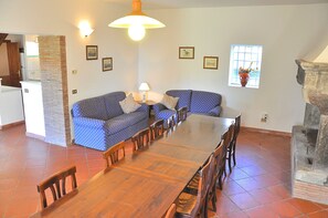 A dining area with a wooden table and chairs, a blue patterned sofa, a brick pillar, and a window with a vase. - Villa i Noccioli   18 sleeps, swimmingpool, estate (CAPRANICA)
