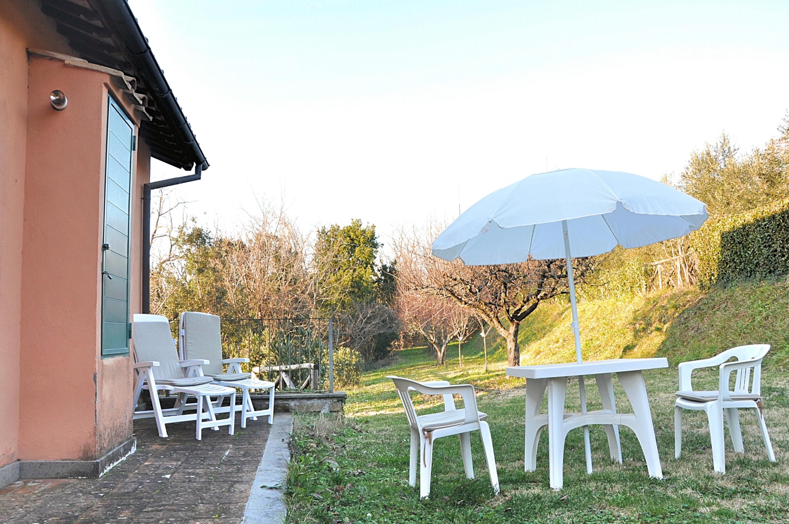A patio with white chairs and a table, an umbrella, and a building with a window.
