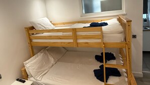 Une chambre avec un lit superposé, une fenêtre située au-dessus du lit du haut et une cuisine visible au fond.