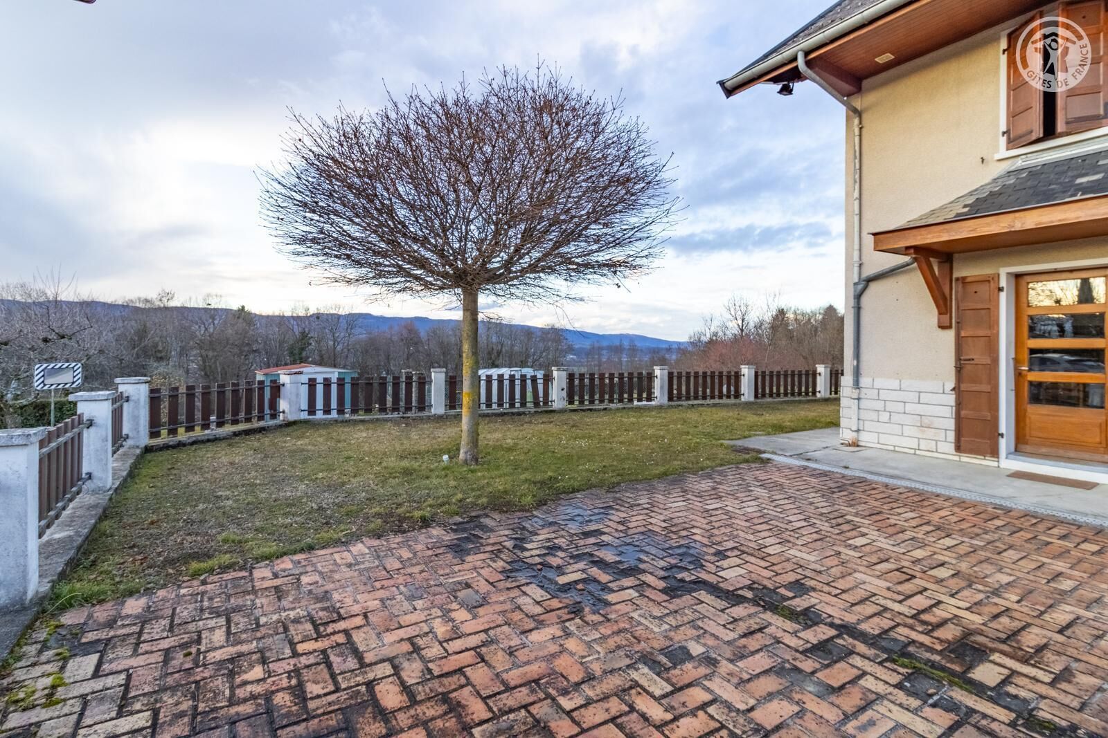A building with a brick pathway leading to a wooden door, surrounded by a fenced area with a tree and mountains in the background.
