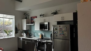A modern kitchen with white cabinets, a stainless steel refrigerator, a microwave, and an oven. There are plants on the countertops and a window with a view of trees. - Cozy studio apartment in charming St. Petersburg perfect for your getaway. (St. Petersburg)