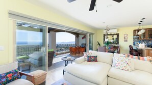 Un vaste espace de vie comprenant un canapé, une table basse et un coin repas avec une table et des chaises. Une grande porte-fenêtre coulissante donne accès à un balcon offrant une vue sur l’océan.