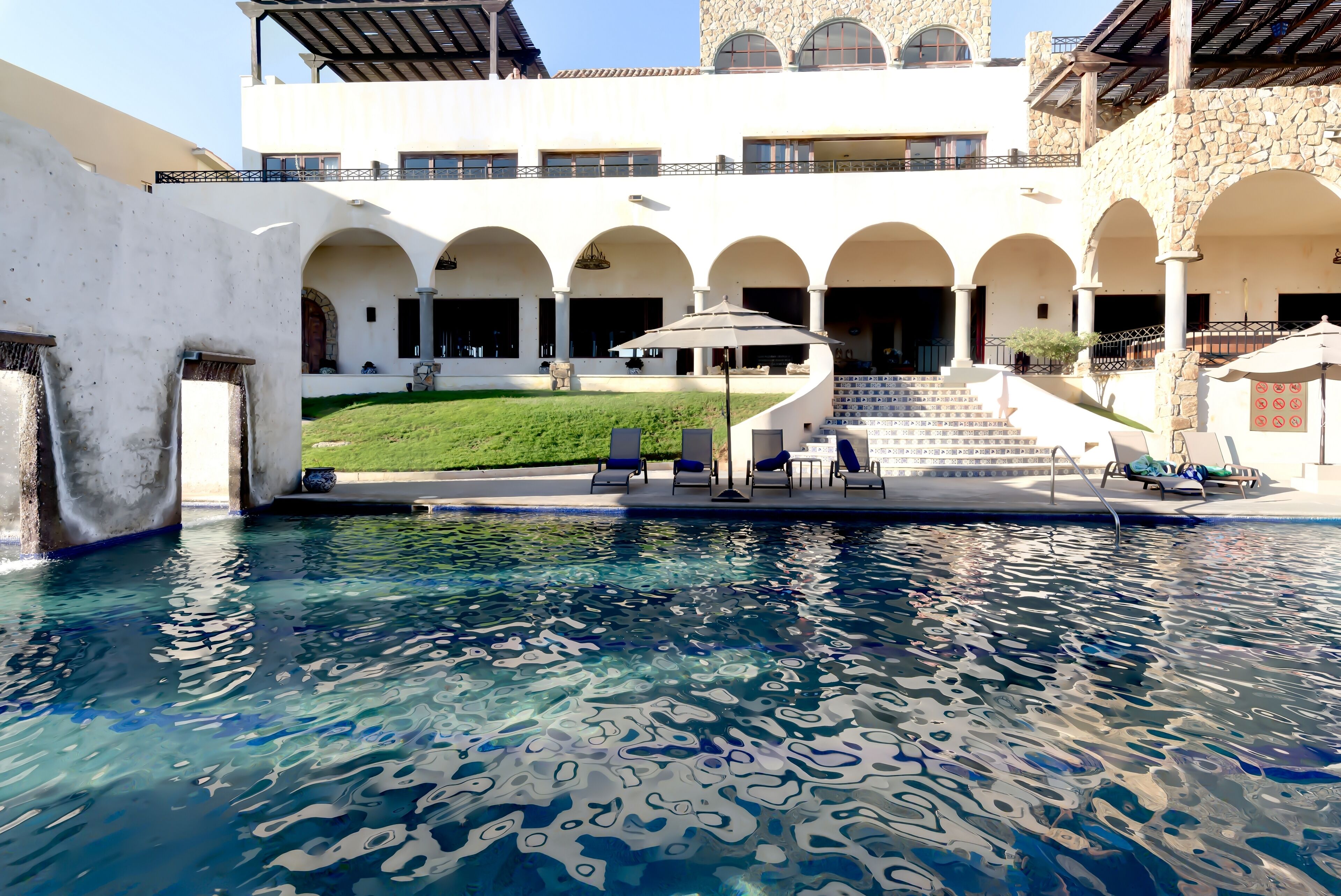 A swimming pool with clear water, surrounded by a building with arched walkways and outdoor seating.