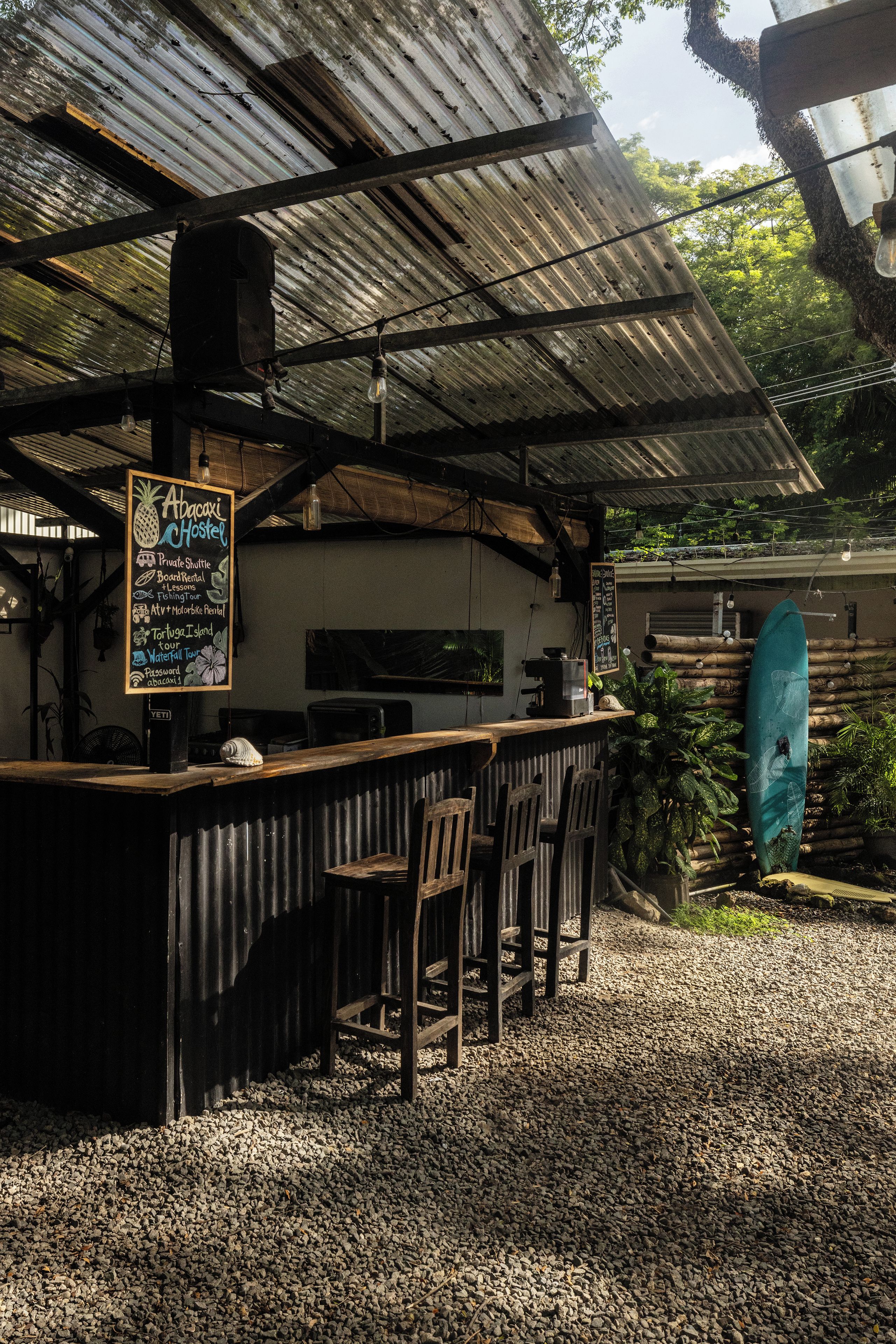 Un área de bar al aire libre con techo de metal rústico, sillas de madera, un menú en una pizarra y una tabla de surf apoyada contra una pared.