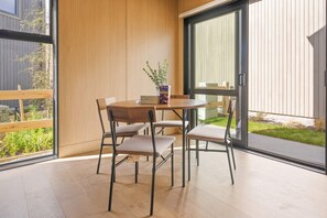 A modern dining area with a round table and four chairs, a vase with a plant, and a view of a garden through large sliding glass doors. - Cynthia's Home away from Home (Queenstown)