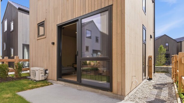 Un edificio residencial moderno con fachada de madera, grandes puertas correderas de vidrio y un espacio exterior visible con un camino de grava y una cerca de madera.