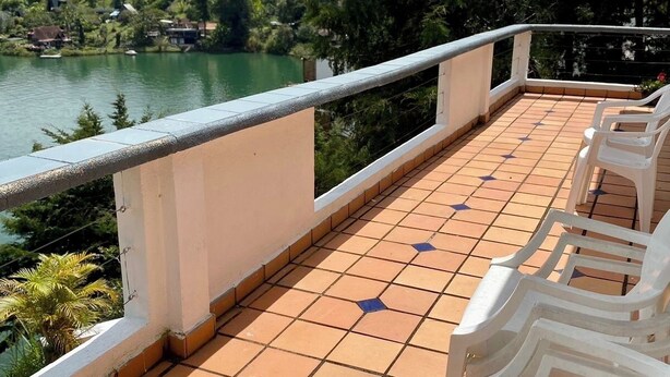 Un balcone con pavimento piastrellato e sedie di plastica bianca, con vista su un lago e sulla vegetazione.