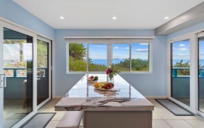 Cozinha moderna com bancada de mármore, uma tigela de fruta e vista para o mar através de grandes janelas.