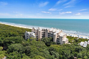 Un hotel costero con varios pisos, rodeado de abundante vegetación y vistas a una playa bajo un cielo azul despejado.