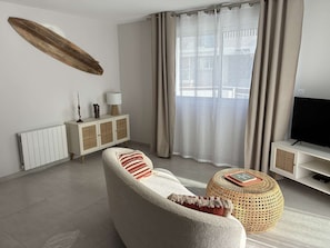 Ein modernes Wohnzimmer mit einer Couch, einem runden Hocker, einem Fernseher, einem Schrank und einem großen Fenster mit Vorhängen.