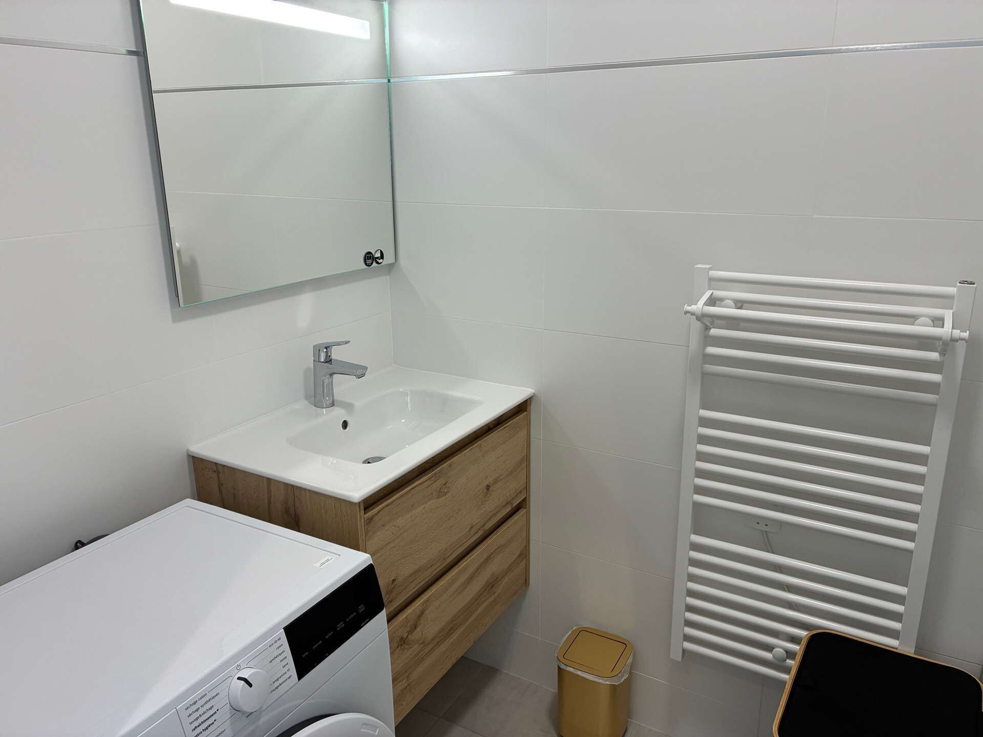 Un bany modern amb un vàter blanc, un tocador de fusta, un mirall, un radiador blanc i una màquina de rentar.