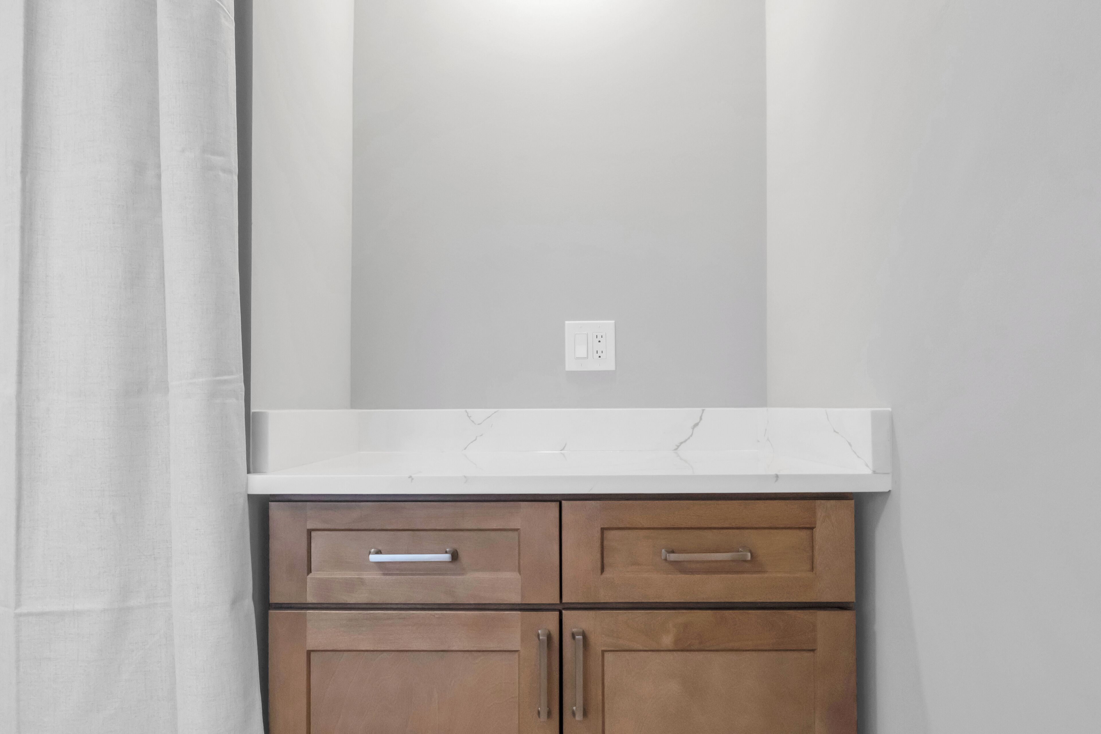 A bathroom with a white countertop, wooden cabinets, and a white wall.