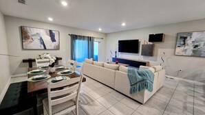 A modern living room with a sofa, a dining table set with plates, and a TV mounted on the wall. - Luxury Disney Home w/ Resort Pools, Tennis & Gym – Near Disney (Kissimmee)