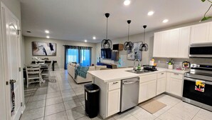 A modern kitchen with white cabinets, a dishwasher, and a large island. There is a dining area with a table and chairs, and a living room with a sofa and a TV. - Luxury Disney Home w/ Resort Pools, Tennis & Gym – Near Disney (Kissimmee)