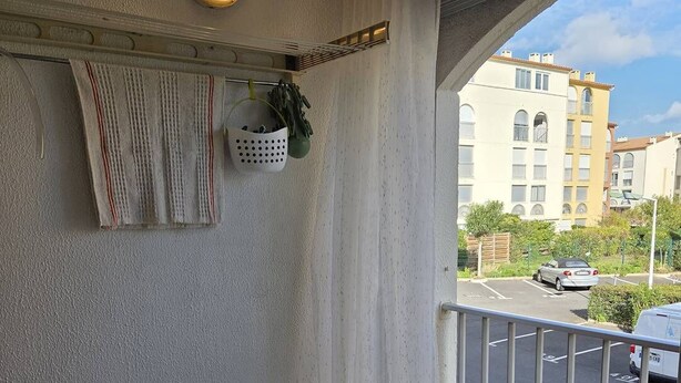 A balcony with a wooden table, chairs, a hanging plant, and a view of a parking lot and buildings. - Lovely 2-room apartment for 4 people in Mt St Martin near the port of Cap d'Agde (LE CAP D AGDE)