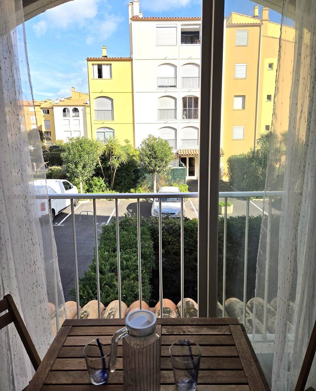 Ein Balkon mit einem Holztisch, zwei Gläsern und einer Kanne. Im Hintergrund sind gelbe Gebäude und ein Parkplatz mit Autos zu sehen.