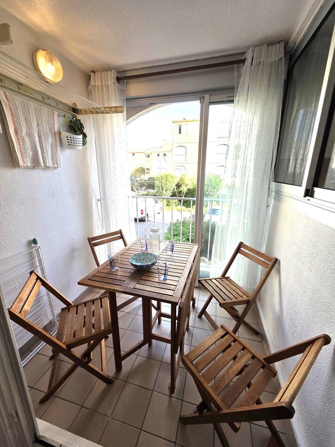 En balkon med et træbord og stole, udsigt til bygninger, og et vindue med gardiner.