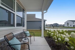 Eine überdachte Terrasse mit zwei Korbstühlen und einem kleinen Tisch, ein Garten mit weißen Blumen und ein Wohngebäude im Hintergrund.
