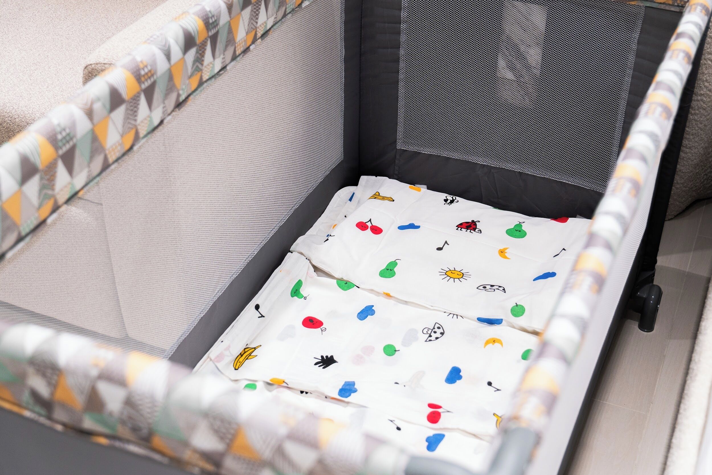 A crib with a white mattress featuring colorful fruit and vegetable prints, surrounded by a grey mesh and a patterned border.