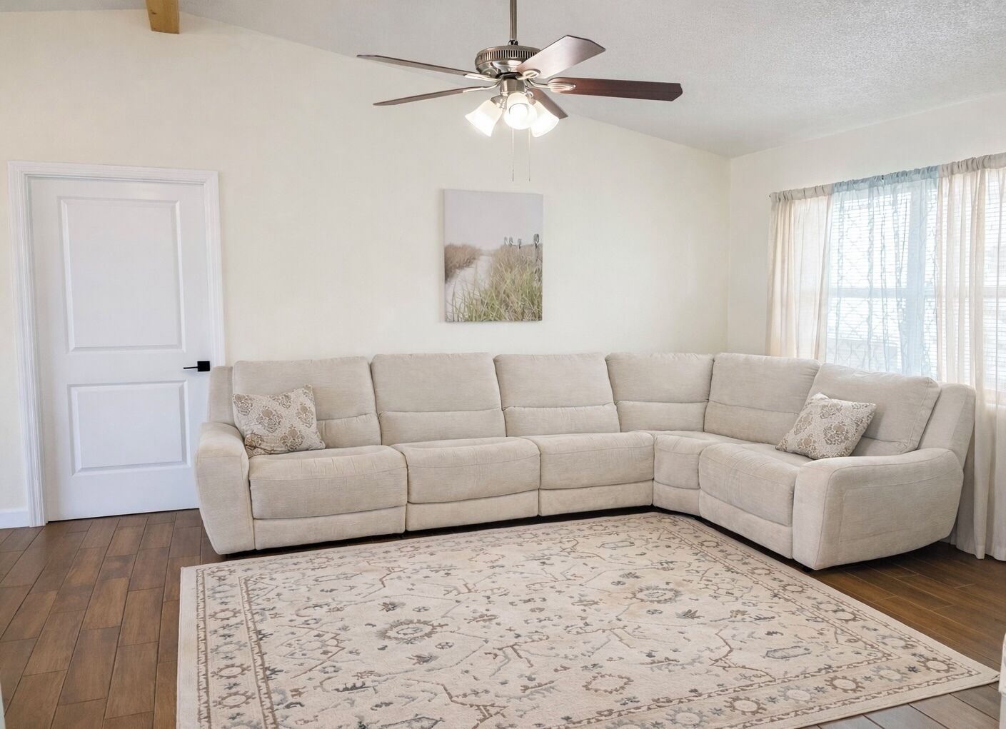 A living room with a beige sectional sofa, a patterned rug, a ceiling fan, and a window with sheer curtains.