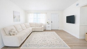 Un soggiorno moderno con un divano angolare beige, un tappeto decorato, un piccolo tavolino bianco e una TV a schermo piatto montata a parete.
